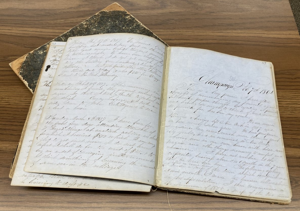 Photograph of two diaries from the 1800s. One diary is open at an angle and stacked upon the other. The open diary has Champaign, Ill. Feb. 7th, 1864 written at the top in large cursive script.