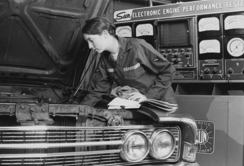 Female mechanic inspects car engine, automotive maintenance 1976