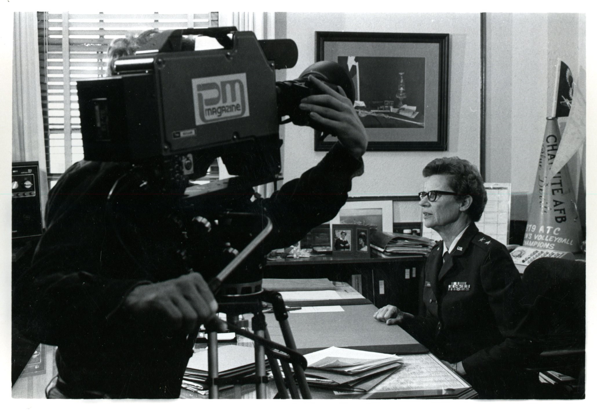 Image of Commander Norma E. Brown in profile, seated at her desk at Chanute. A video camera is positioned in front of her.