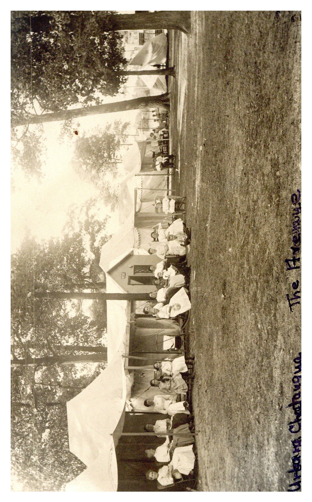 Canvas tents lined up in a wooded clearing with a row of women and children seated in chairs in front of the tents.  