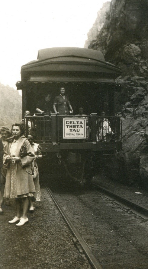 Black and white photograph of a train caboose next to mountains. The train is stopped and women are standing outside, around the caboose.