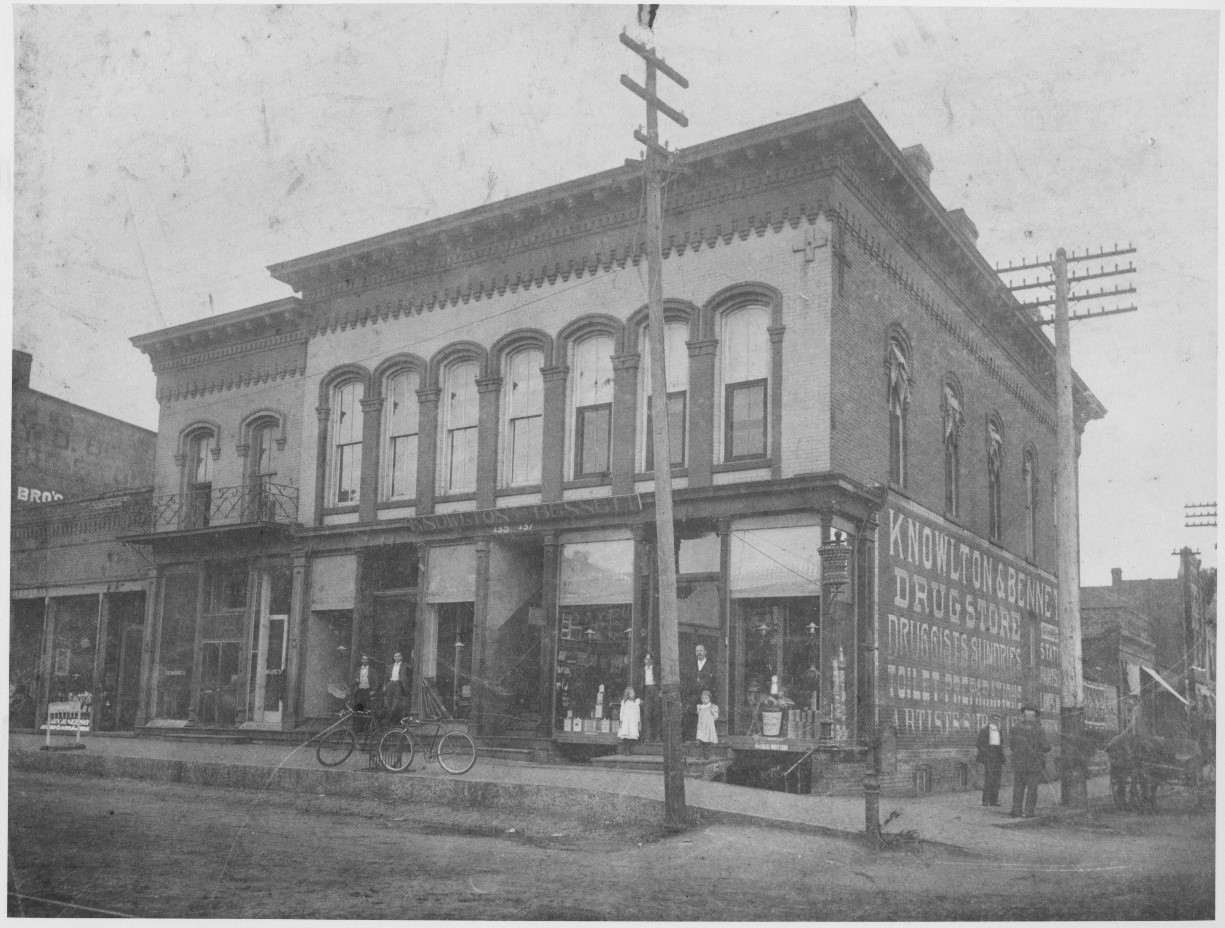 A black and white photograph of a two story building taken in 1871. A few people are visible standing outside the building, some with bicycles, and one with a horse.