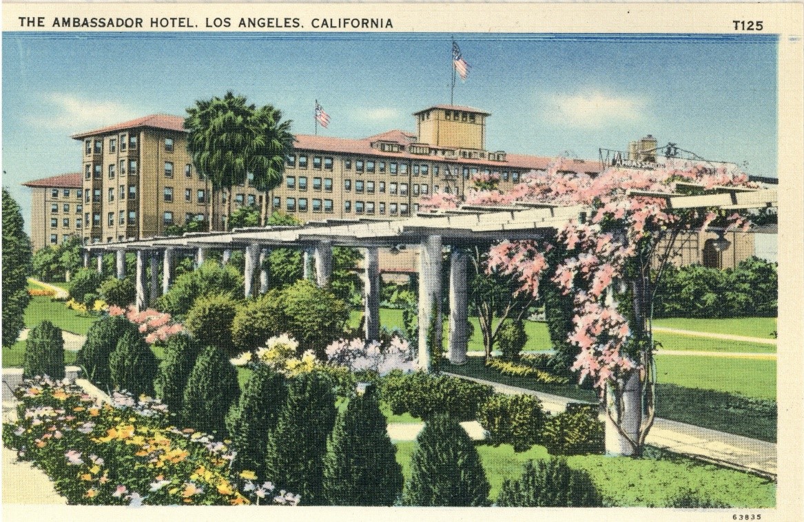 Color postcard of The Ambassador Hotel in Los Angeles. White trellises are surrounded by plants and flowers with the hotel in the background.