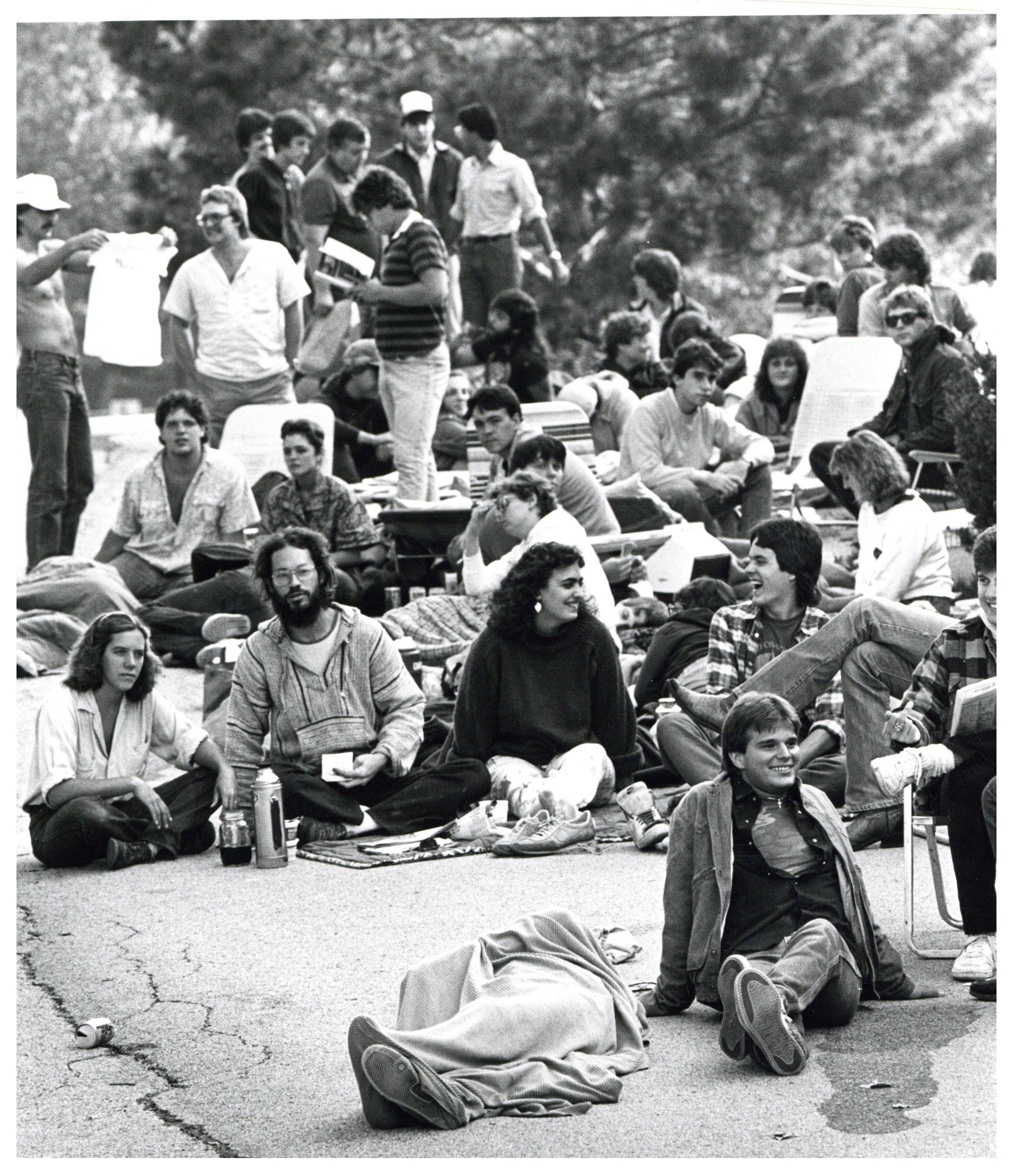 Black and white photograph showing would-be concert goers waiting to buy tickets for Farm Aid. They are seated on the ground, laying down, and standing.