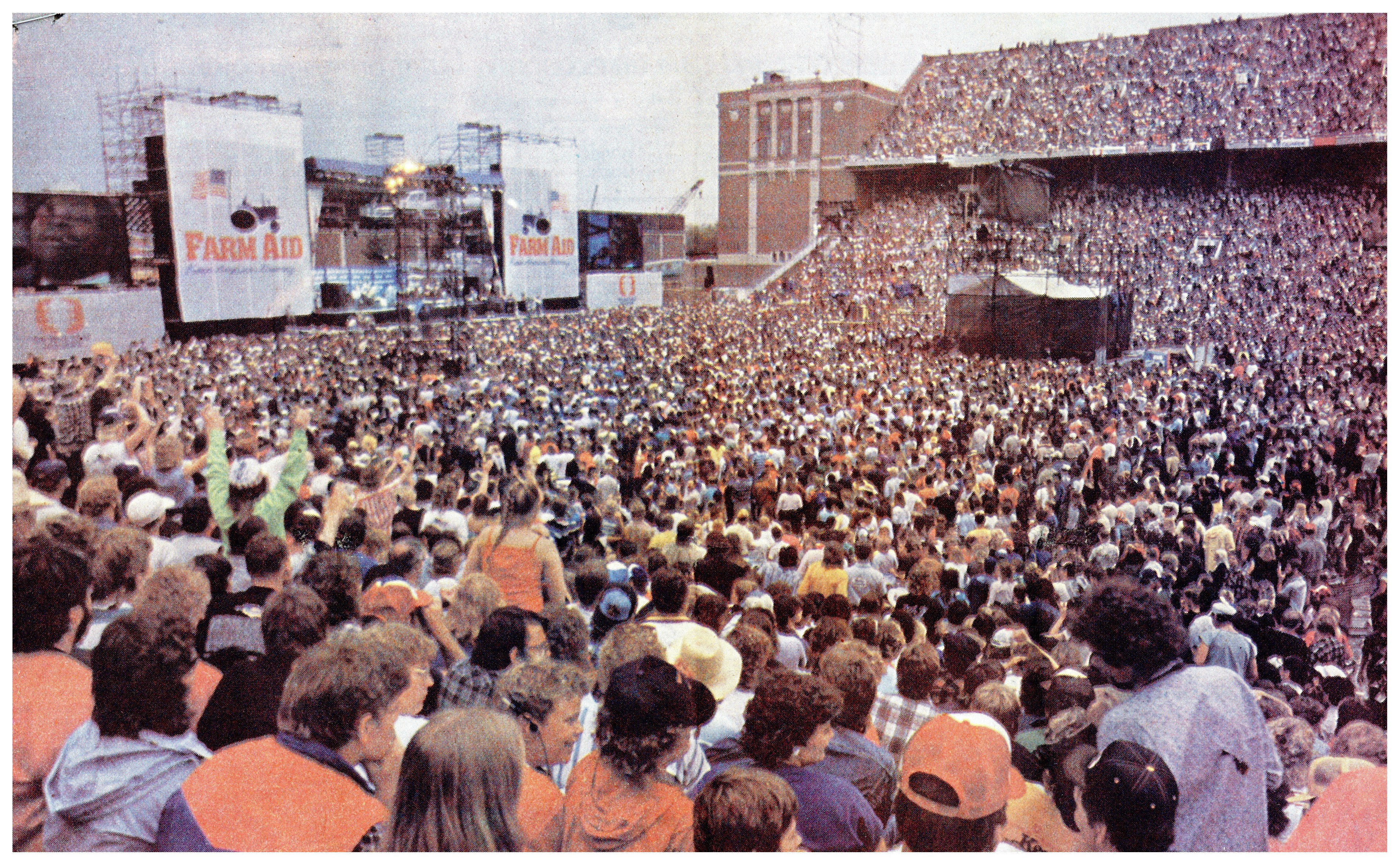 A color photograph of the crowd and stage at Farm Aid in Memorial Stadium. The field and stands are packed with concert-goers.