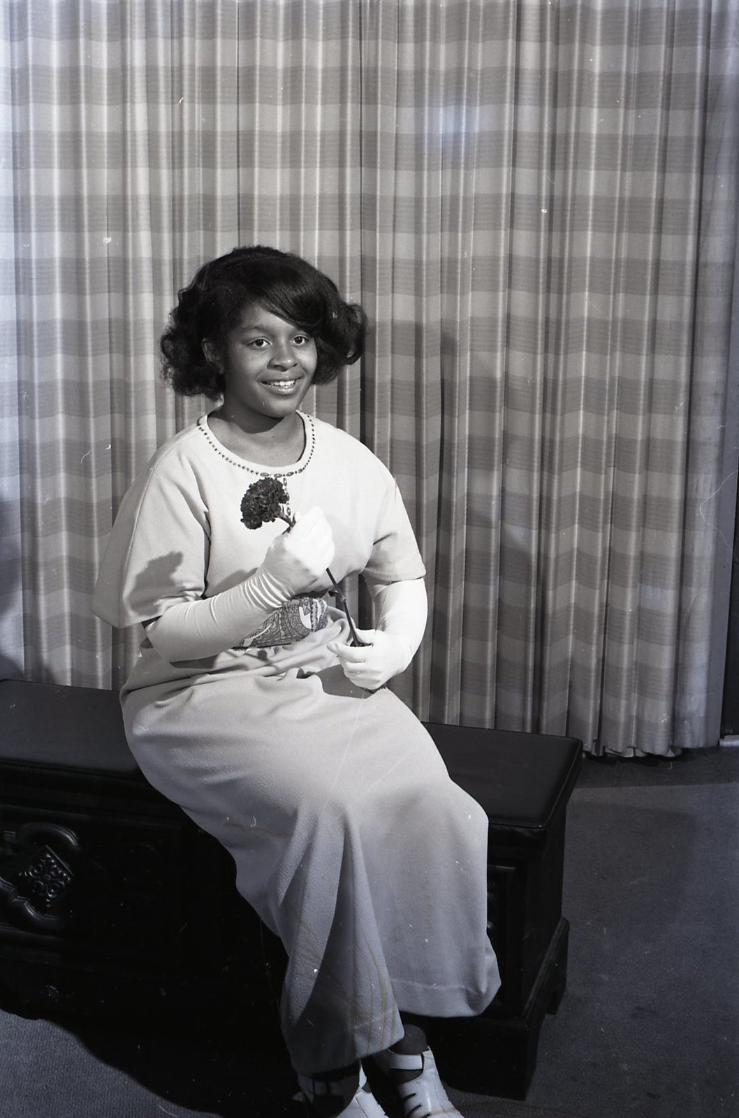 Black and white photograph of a young African American woman in a white dress, seated, holding a flower and smiling at the camera.