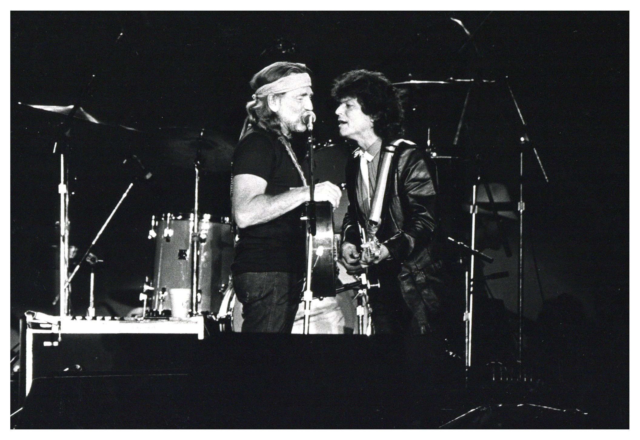Black and white photograph of Willie Nelson and Mick Jagger singing into the same microphone on stage at Farm Aid. They are both holding guitars. A drum kit is visible behind them.