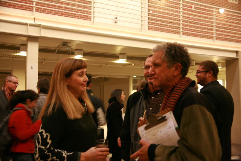 A man and a woman stand facing each other in conversation. Other people are visible in the background.