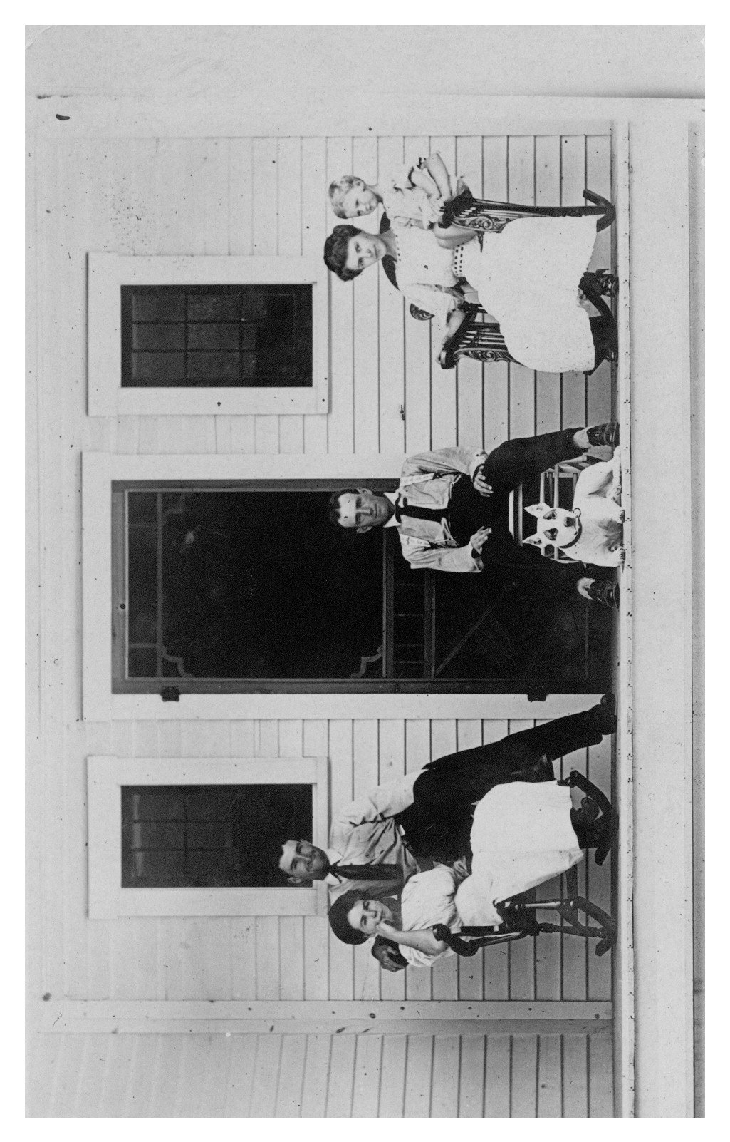 Four people sitting on the front porch of a white frame house. On the left is a man and a woman sharing a rocking chair, in the middle is a seated man with a dog sitting in front of him, to the right is a woman and a small child sharing a rocking chair.  in fr