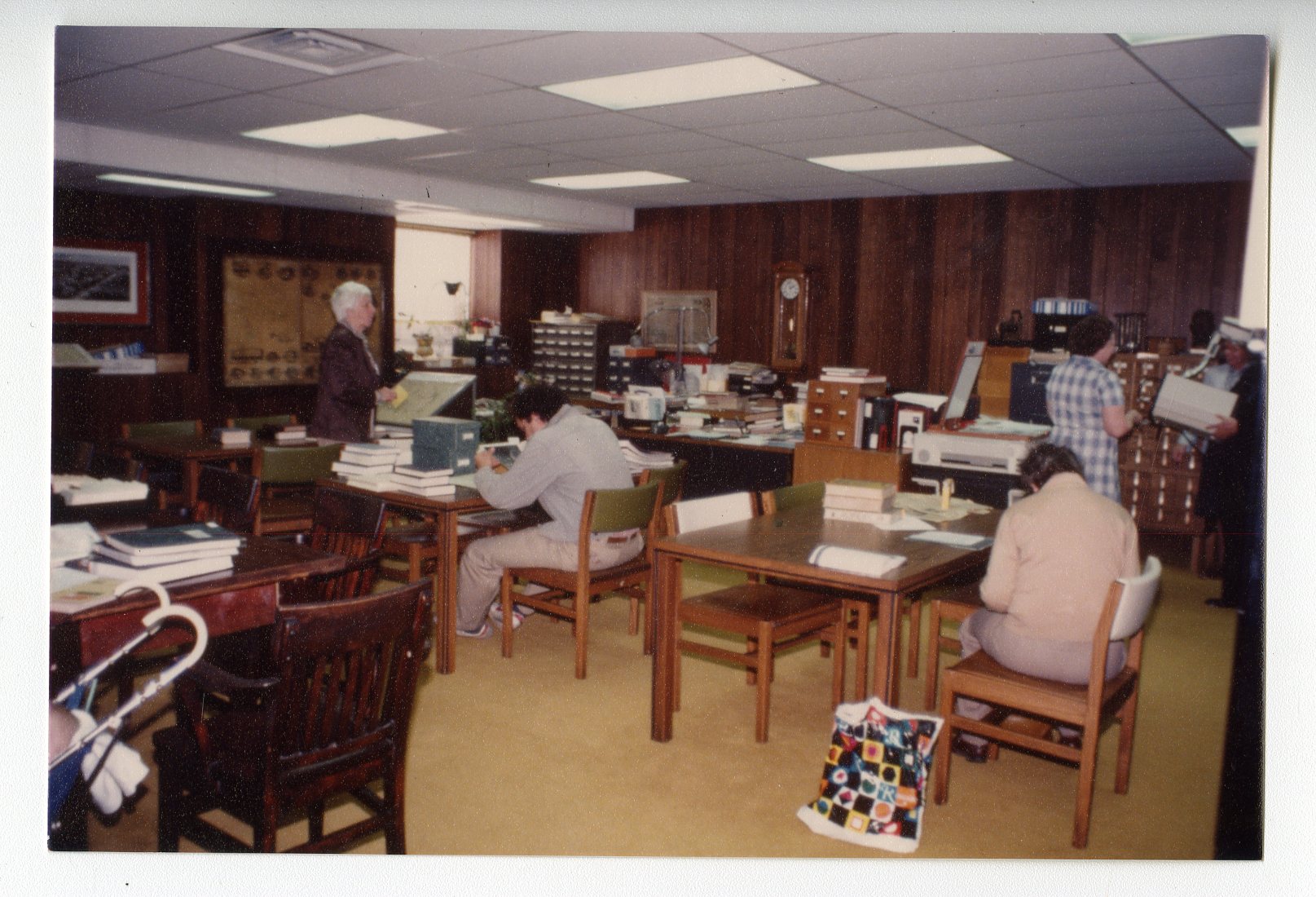 A basement room with wood paneled walls houses many card catalogs and reading tables. Researchers are seated at the tables looking at books.