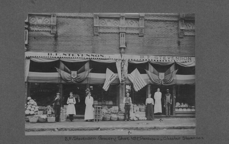 Black and white photograph of the front of a grocery store in the early 1900s. Flags are hanging above the doors, and piles and baskets of fruit are arranged outside. Banners above the store read, "B.F. Stevenson." A group of people is standing outside. The center figure is B.F. Stevenson, his son, Chester is second from the right. The woman in the middle is possible Lucia Stevenson.