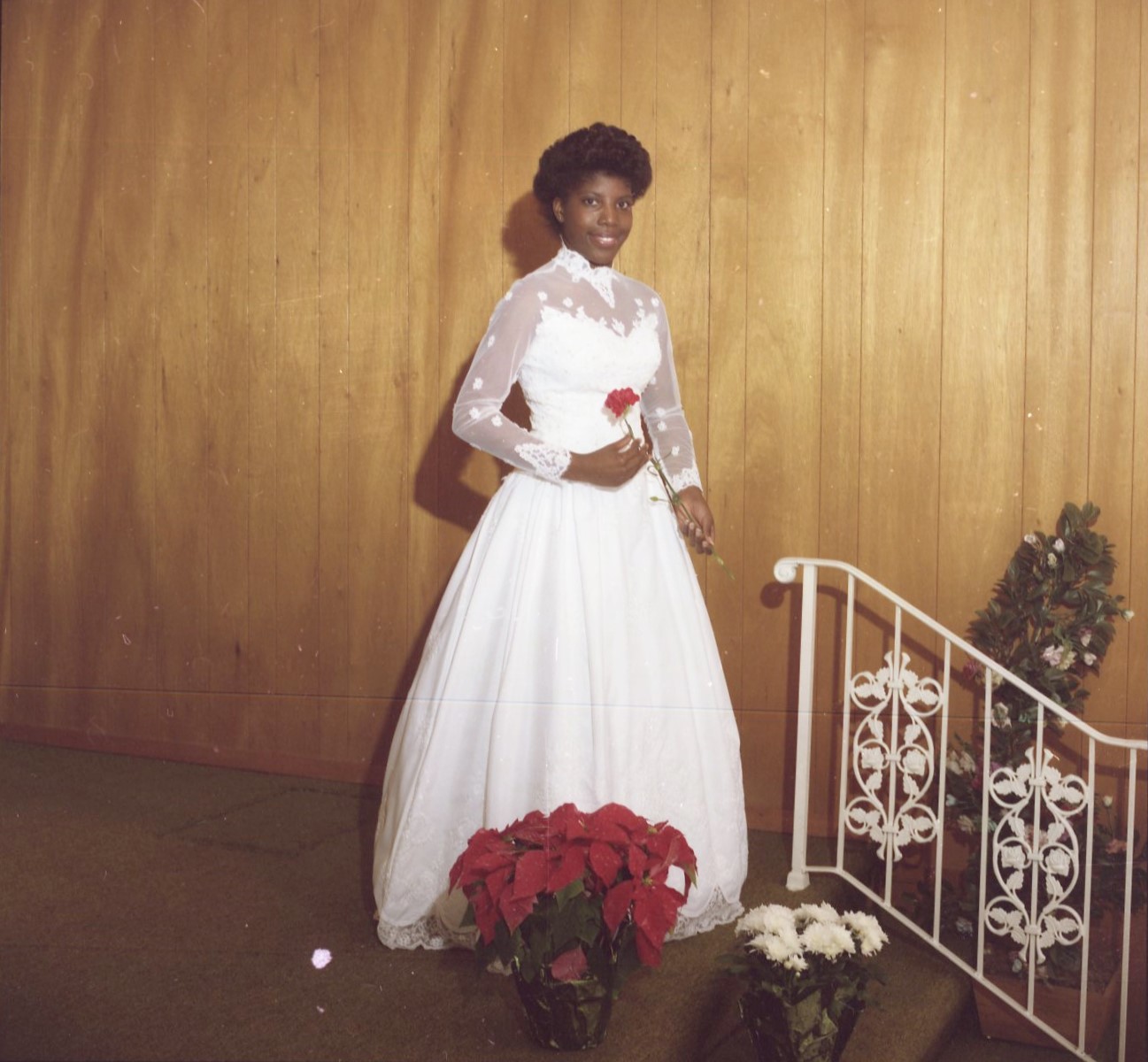 A color photograph of an African American young woman in a white wedding dress. She is holding a red rose. She is a debutante at the 1986 Cotillion Ball.