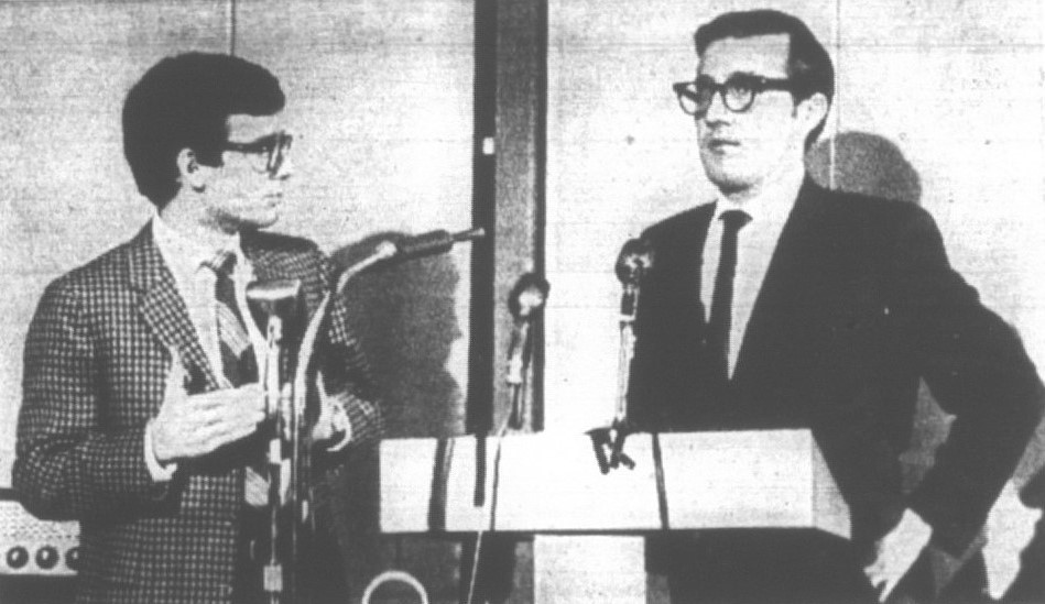Photograph from a newspaper of two men standing in front of a podium with microphones.
