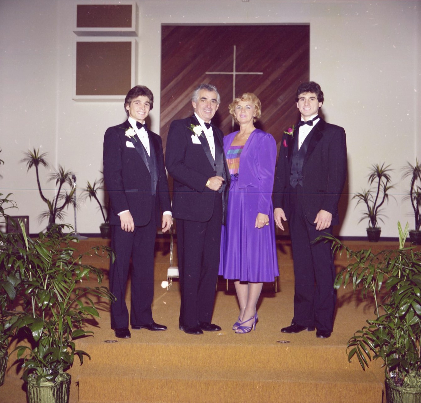 A color photograph of three men in black tuxedos and a woman in a purple dress. The are standing at the altar of a church, posing for a photograph at a wedding.
