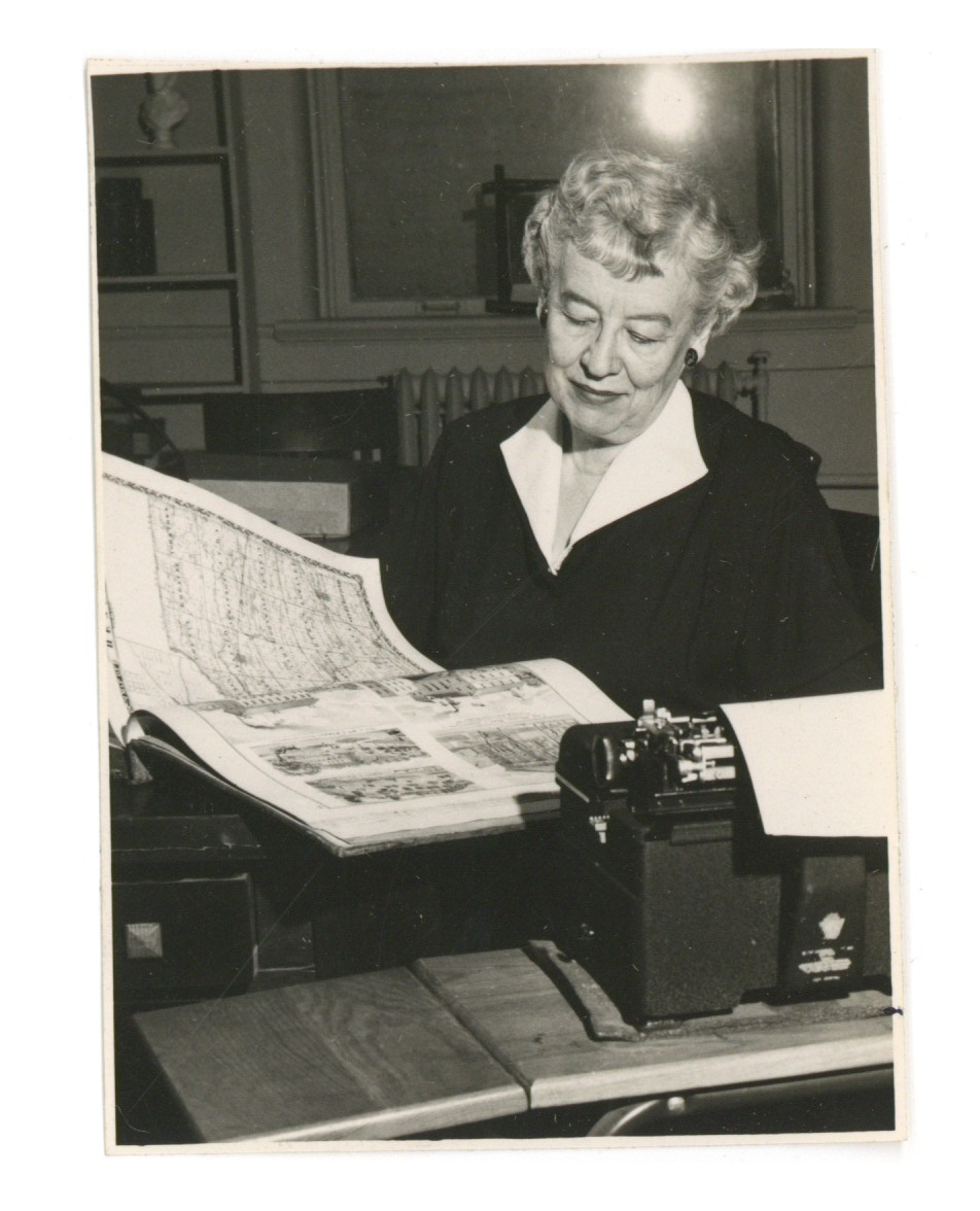 Black and white photograph of an older woman seated as a desk. She is looking at an open atlas in front of her. There is a typewriter on the desk.