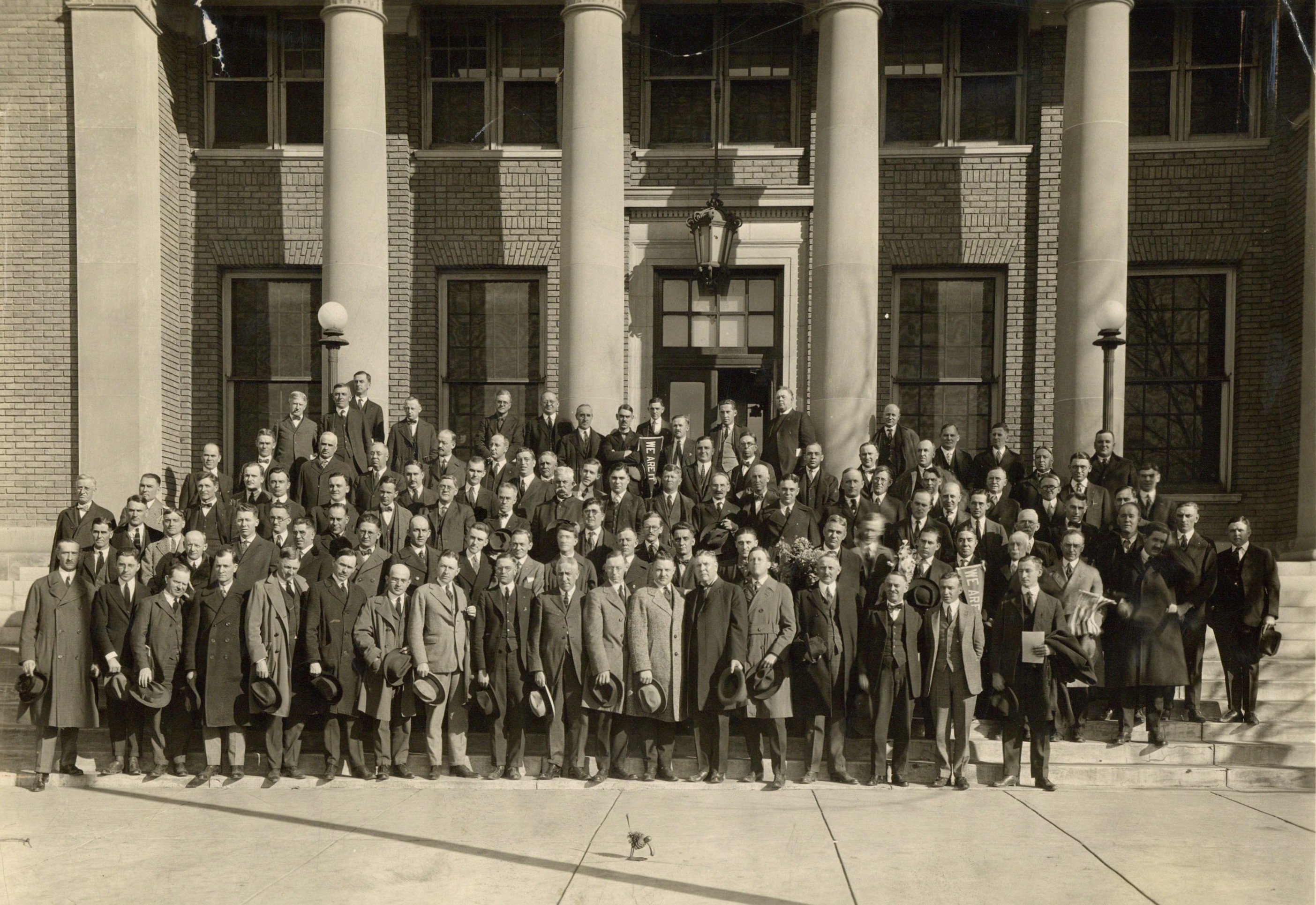 Black and white photograph of men lined up and standing on stairs in front of a building with columns. 