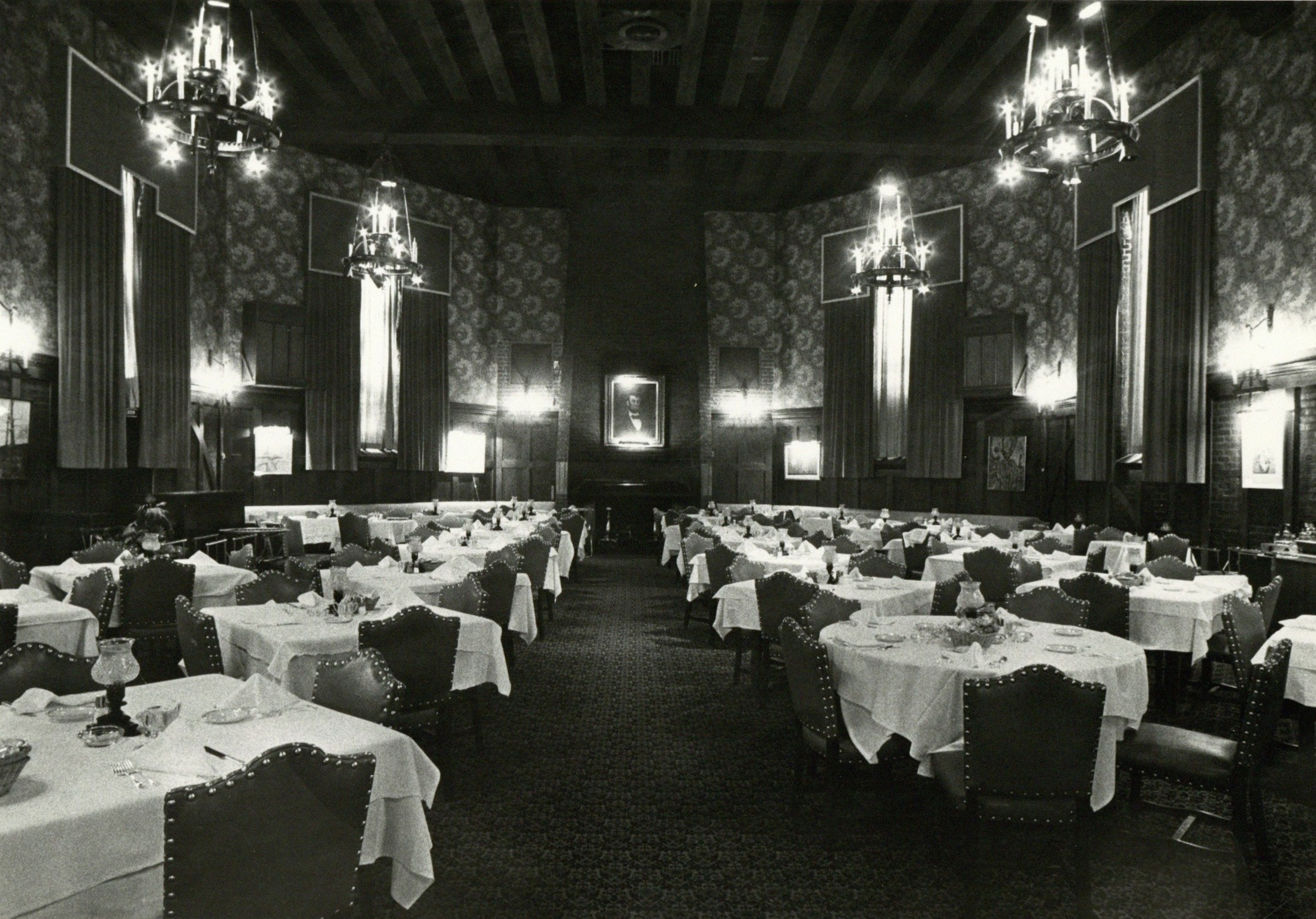 Black and white photograph of the Luncheon Room in the Urbana Lincoln Hotel taken in 1975. The room is full of empty tables with white table cloths, patterns wallpaper, long drapes on the windows and chandeliers.