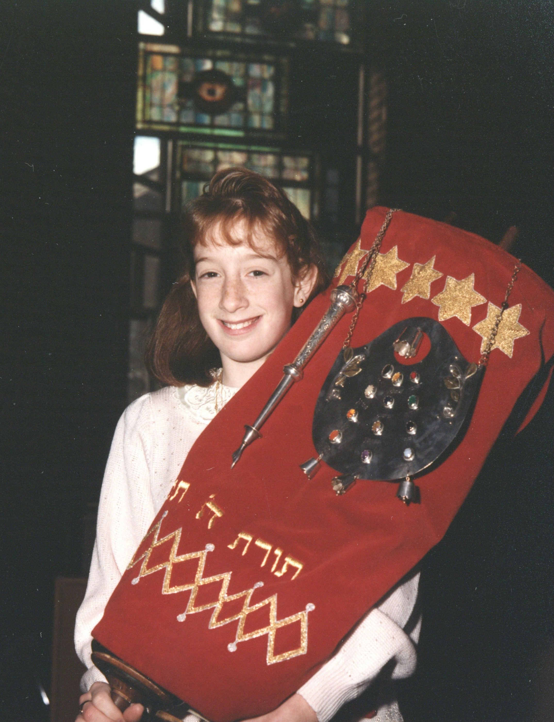 A color photograph of a young girl at her bat mitzvah. She is holding the torah and smiling.
