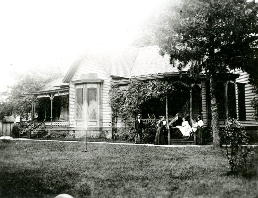 Black and white photograph of 5 people socializing on the porch of a house.