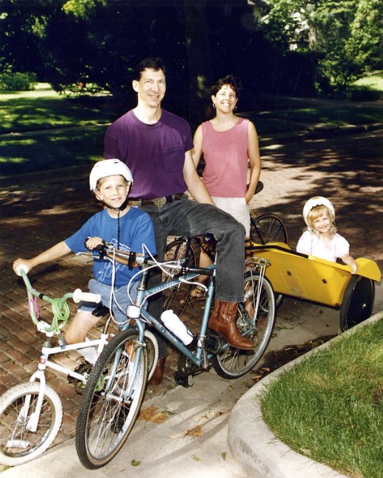 A color photograph of two adults and two children on bikes. The smallest child is sitting in a bike trailer.