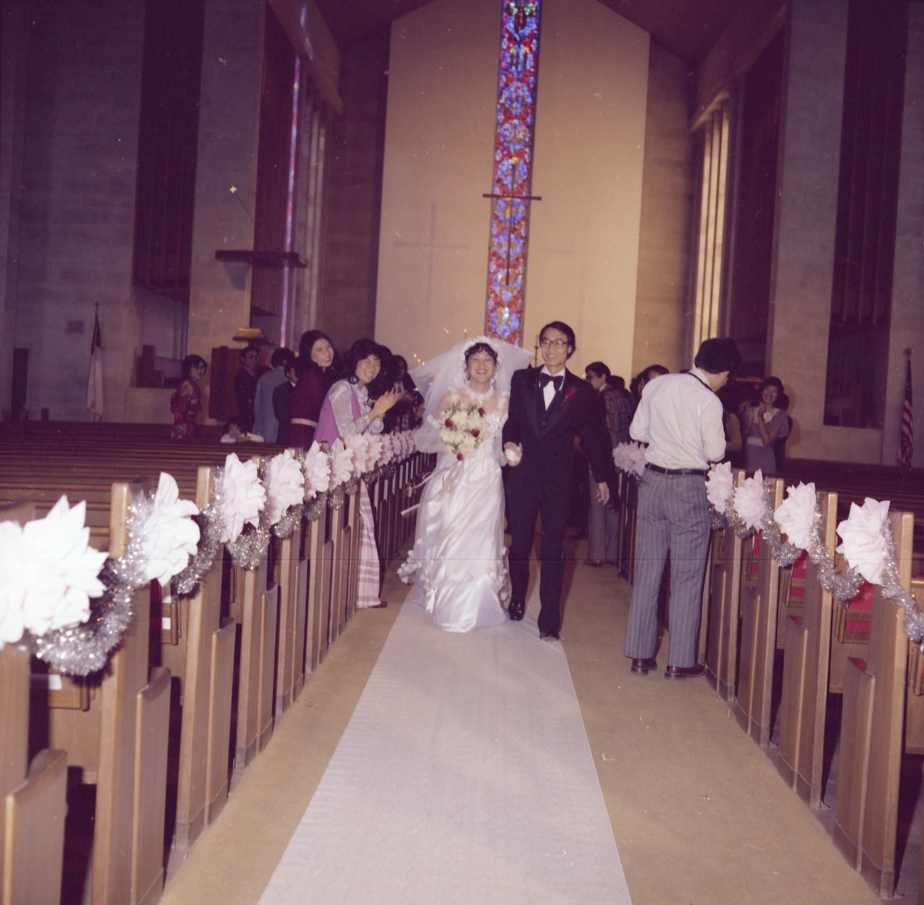 A color photograph of a newlywed Asian couple walking down the aisle of a church after their wedding ceremony.