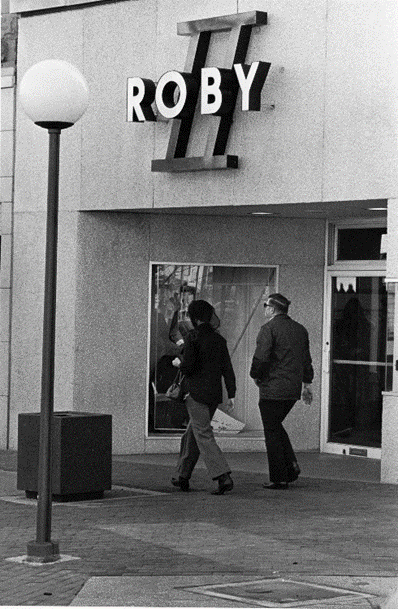 Black and white photograph of two people walking past a storefront. The sign above the door reads, Roby II. 