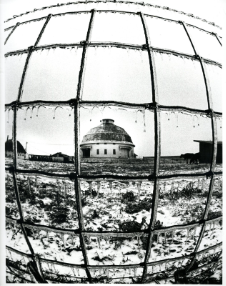 A black and white photograph of a round barn taken through fence wires. There is snow on the ground.