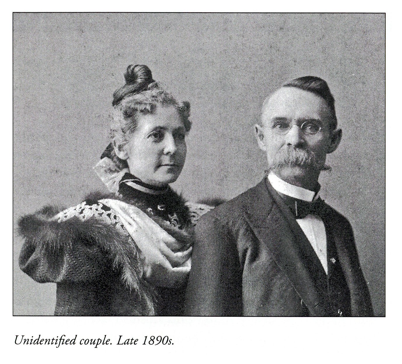 Black and white image of a man and woman. The women has an elaborate bun on top of her head and is wearing a dress with puffy, fur and lace-trimmed sleeves.