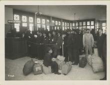 Immigrants with baggage lined up at teller's windows marked money exchange